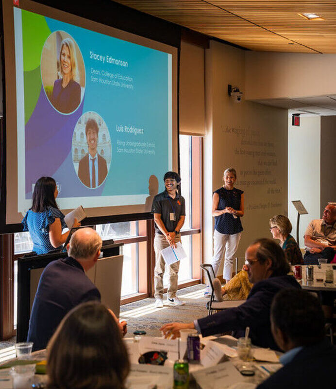 Stacey Edmonson, a teacher-candidate, and a DFI staff member standing next to a projector screen with event attendees sitting in the background